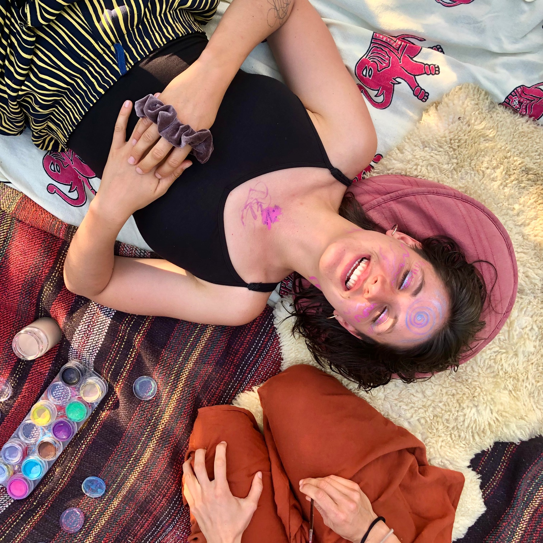 A laughing woman with face paint lying on colourful blankets